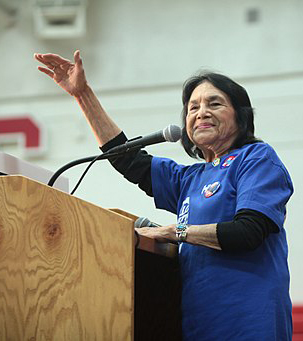 A photo of activist Dolores Huerta in a blue shirt with her arm raised, speaking at a rally.